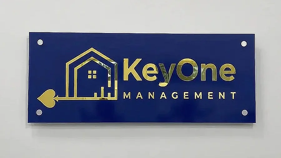 A blue KeyOne Management panel in gold lettering, held off a white wall on four stainless standoffs
